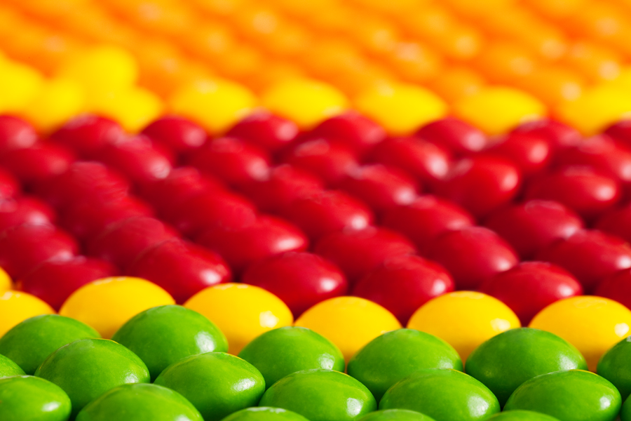 This Brian Charles Steel Photograph is composed of round candy in various colors. The candies are arranged in horizontal rows of the same color. The bottom portion of the frame has two rows of green candy. A single row of yellow candy borders the green candy. Behind the yellow candy are rows of red candy, and there is a row of yellow candy on the side. The top portion is filled with orange candy.  